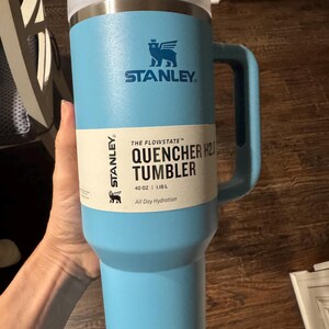 May include: A light blue Stanley Flowstate Quencher H2.0 tumbler with a handle and a clear lid. The product label reads: Stanley The Flowstate Quencher H2.0 Tumbler 40 oz | 1.18 L All Day Hydration. The cup features a winged bear logo in dark blue.