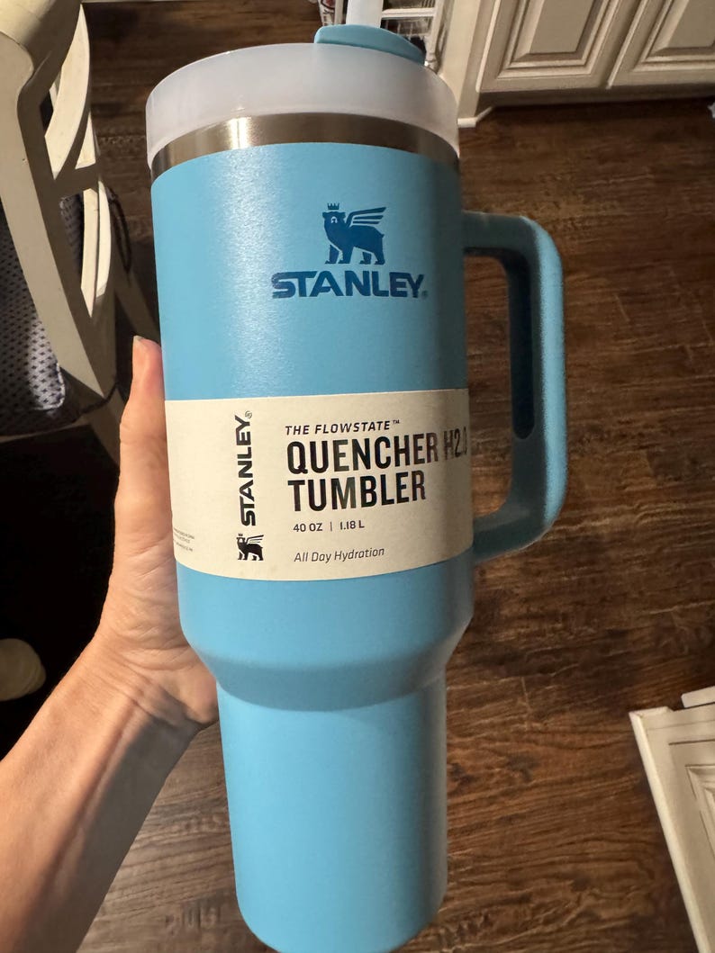 May include: A light blue Stanley Flowstate Quencher H2.0 tumbler with a handle and a clear lid. The product label reads: Stanley The Flowstate Quencher H2.0 Tumbler 40 oz | 1.18 L All Day Hydration. The cup features a winged bear logo in dark blue.