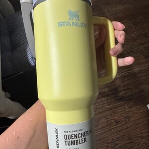 May include: A pale yellow Stanley Quencher H2.0 Flowstate tumbler with a 1.18 L capacity. The cup features a large side handle, a stainless steel rim, and a translucent lid. A product label on the lower section displays the text Stanley The Flowstate Quencher H2.0 Tumbler 40 oz 1.18 L All Day Hydration.