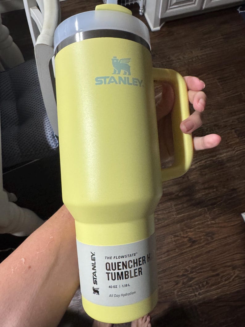May include: A pale yellow Stanley Quencher H2.0 Flowstate tumbler with a 1.18 L capacity. The cup features a large side handle, a stainless steel rim, and a translucent lid. A product label on the lower section displays the text Stanley The Flowstate Quencher H2.0 Tumbler 40 oz 1.18 L All Day Hydration.