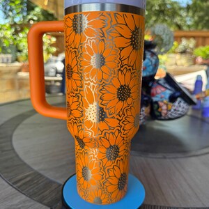May include: A bright orange insulated tumbler with a handle, featuring a repeating sunflower pattern in black outlines. The cup has a white rim, an orange lid, and a clear straw. It stands on a circular blue and black base for display purposes.