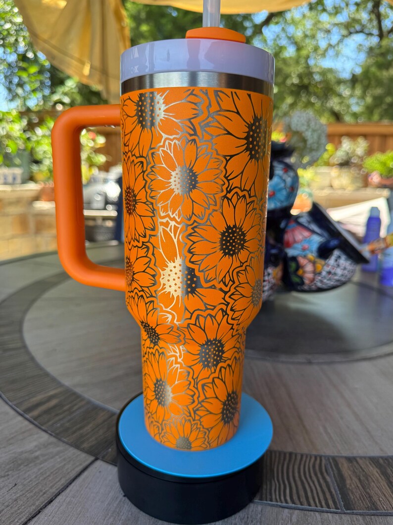 May include: A bright orange insulated tumbler with a handle, featuring a repeating sunflower pattern in black outlines. The cup has a white rim, an orange lid, and a clear straw. It stands on a circular blue and black base for display purposes.
