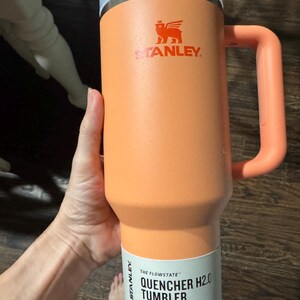 May include: A peach-coloured Stanley Quencher H2.0 Flowstate tumbler with a 1.18 L capacity. The insulated cup features a matching handle, a translucent lid, and a white product label printed with the text The Flowstate Quencher H2.0 Tumbler 1.18 L.
