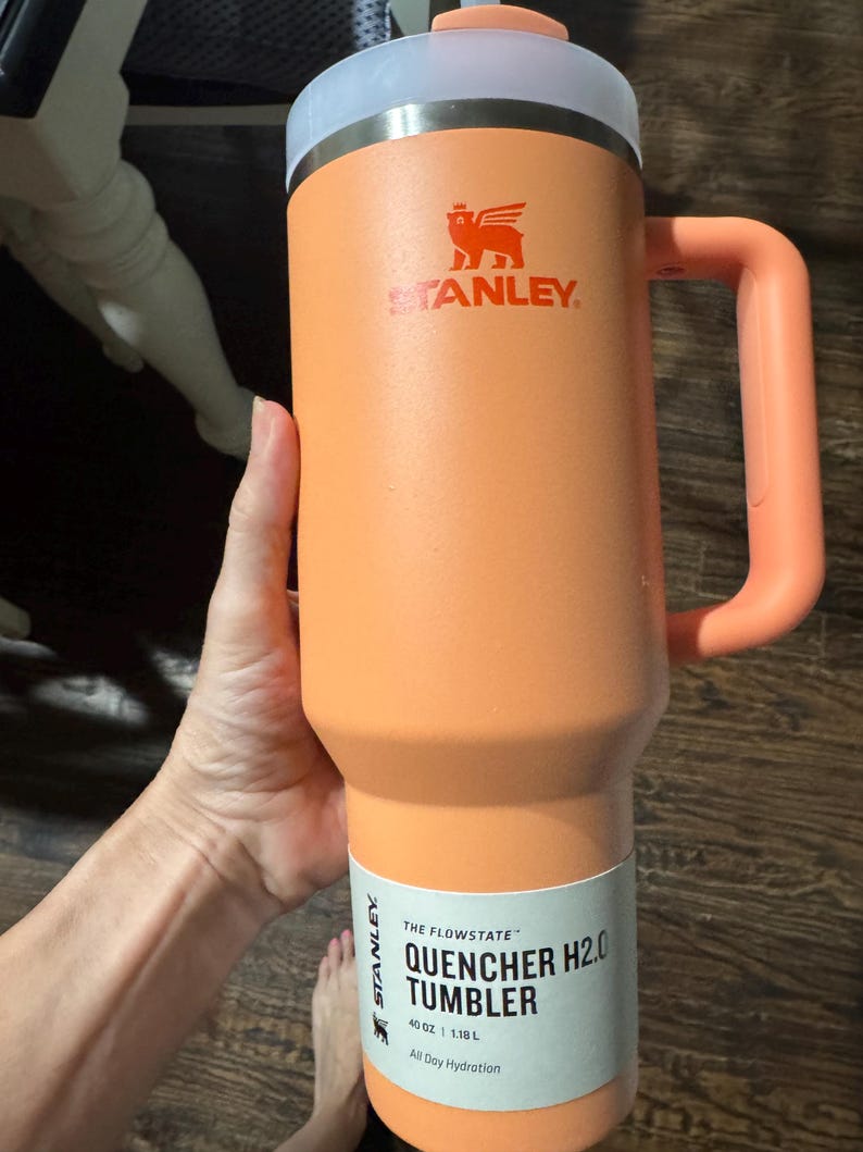 May include: A peach-coloured Stanley Quencher H2.0 Flowstate tumbler with a 1.18 L capacity. The insulated cup features a matching handle, a translucent lid, and a white product label printed with the text The Flowstate Quencher H2.0 Tumbler 1.18 L.