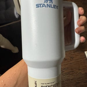 May include: A white Stanley Quencher H2.0 FlowState tumbler with a 1.18 litre capacity. The insulated cup features a large side handle, a metallic rim at the top, and a blue bear logo. A beige product label at the base displays the text 40 OZ | 1.18 L.