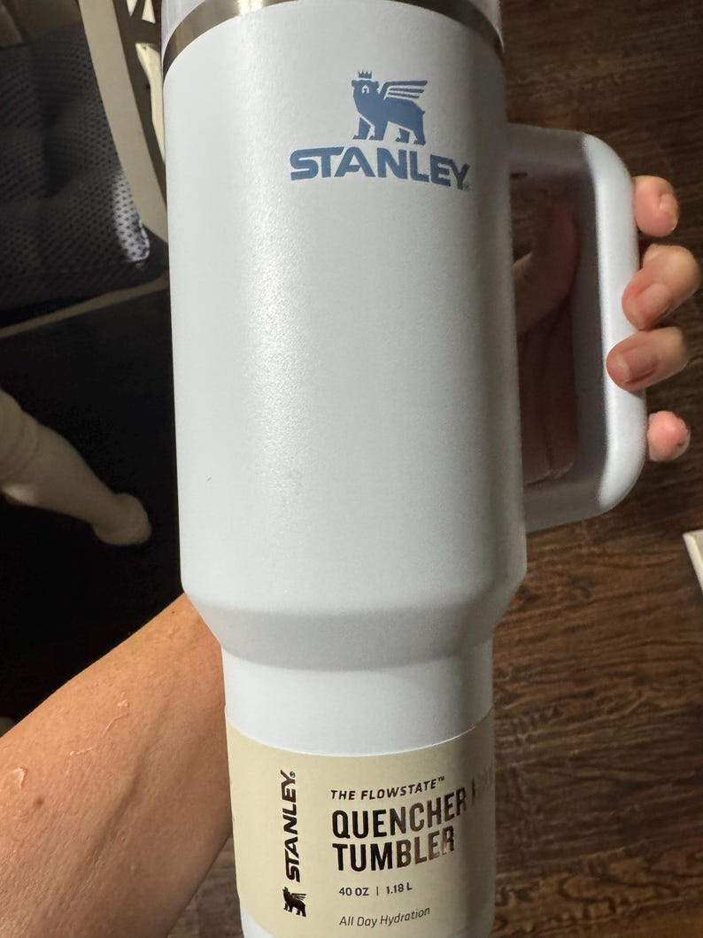 May include: A white Stanley Quencher H2.0 FlowState tumbler with a 1.18 litre capacity. The insulated cup features a large side handle, a metallic rim at the top, and a blue bear logo. A beige product label at the base displays the text 40 OZ | 1.18 L.
