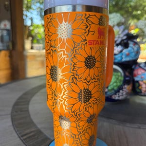 May include: A bright orange Stanley tumbler featuring a repeating sunflower pattern in a metallic silver outline. The cup includes a matching orange handle, a white lid, and a white straw. The Stanley logo is visible in red near the top rim.