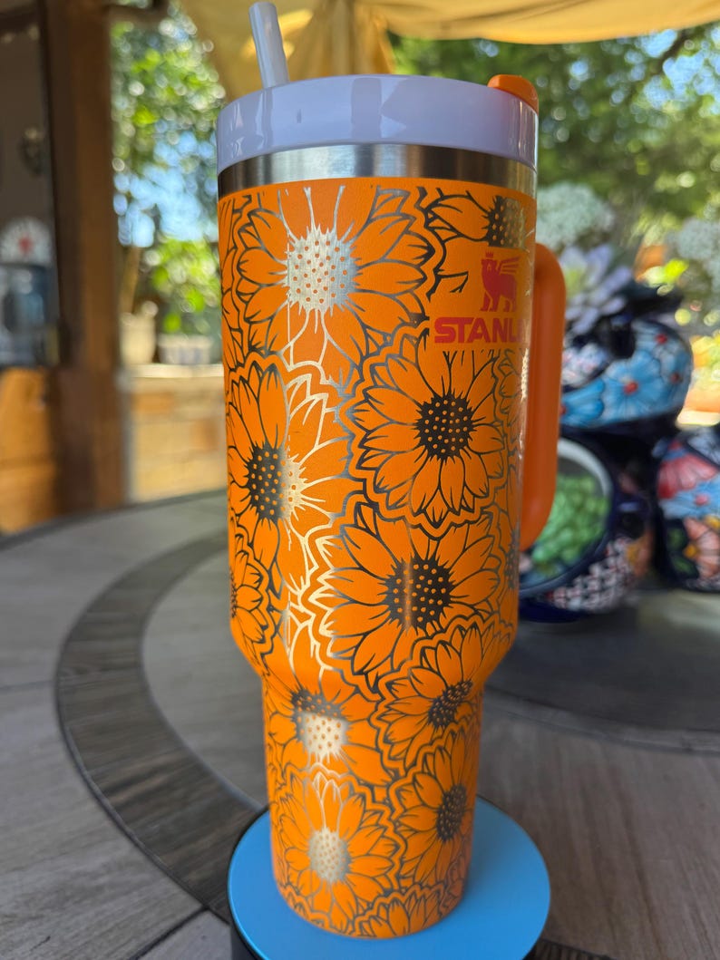 May include: A bright orange Stanley tumbler featuring a repeating sunflower pattern in a metallic silver outline. The cup includes a matching orange handle, a white lid, and a white straw. The Stanley logo is visible in red near the top rim.