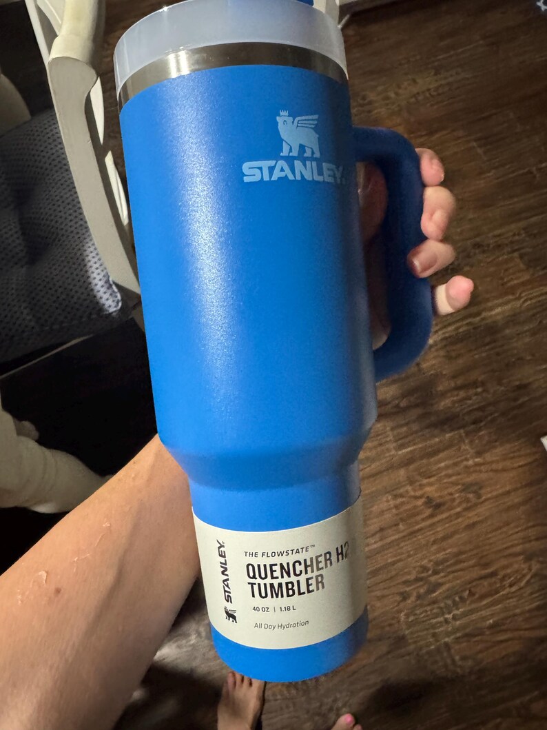 May include: A bright blue Stanley Quencher H2.0 FlowState tumbler with a 1.18 L capacity. The insulated cup features a sturdy side handle, a clear lid, and a white product label printed with the text Stanley The Flowstate Quencher H2.0 Tumbler 1.18 L.