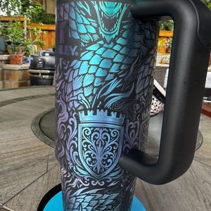 May include: A tall, cylindrical stainless steel tumbler with a black handle and a black lid. The exterior features a vibrant, iridescent blue and purple dragon scale pattern overlaid with a detailed crest design and the Stanley brand logo.
