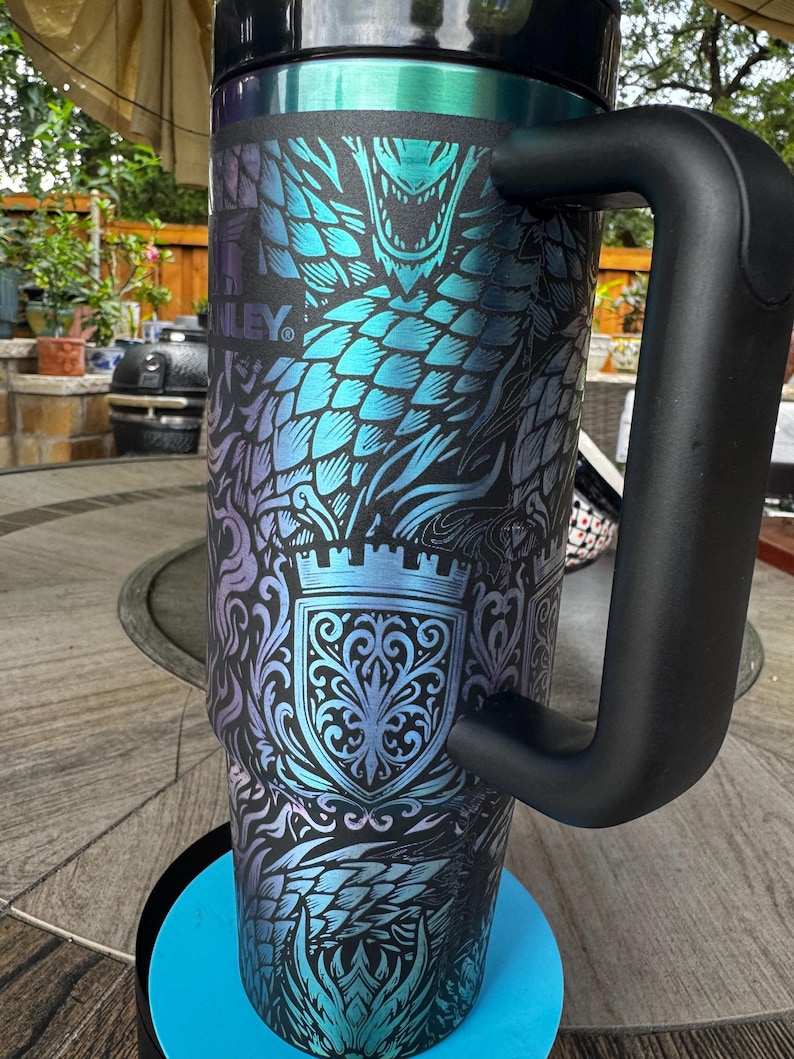 May include: A tall, cylindrical stainless steel tumbler with a black handle and a black lid. The exterior features a vibrant, iridescent blue and purple dragon scale pattern overlaid with a detailed crest design and the Stanley brand logo.