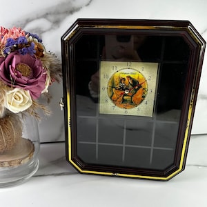 May include: A rectangular, dark brown clock with a gold trim and a glass front. The clock face features a vintage illustration of a couple. The clock is labeled "European Classic".