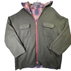 May include: A dark olive green fleece jacket featuring a full-length front zipper with a bright pink trim. The jacket includes two chest pockets with flaps, two lower side pockets, and a hood lined with a multi-colored plaid fabric pattern.