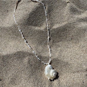 Könnte beinhalten: Eine zarte Halskette mit einem großen, weißen Muschelanhänger. Die Kette ist mit kleinen, weißen Perlen und goldfarbenen Akzenten versehen. Der Schmuck liegt auf Sand und erzeugt eine Strandästhetik.