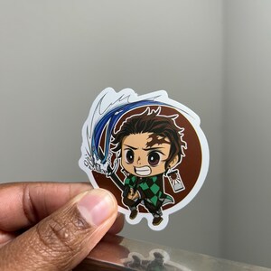 May include: A die-cut vinyl sticker featuring a chibi-style anime character with dark hair, a green and black checkered haori, and a sword with a blue water effect. The character has a scar on the forehead and wears hanafuda-style earrings.