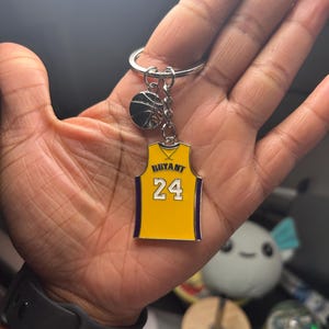 Può includere: Un portachiavi a forma di maglia da basket gialla e viola con il nome "Bryant" e il numero "24" sul davanti. Il portachiavi include anche un piccolo ciondolo a forma di palla da basket.