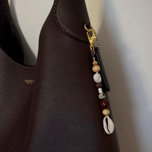 May include: A dark burgundy leather Coach hobo bag with a gold-tone clasp and a beaded charm. The charm features wooden beads, a cowrie shell, and a black rectangular accent.