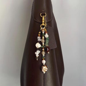 May include: A dark brown leather handbag featuring a decorative beaded charm attached to the side ring. The charm consists of multiple strands of assorted beads in shades of white, green, brown, and clear, including various shapes and textures.