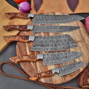 May include: A set of six Damascus steel kitchen knives with wooden handles. The blades have a unique patterned design. The handles are decorated with floral accents. The knives are displayed on a wooden cutting board.