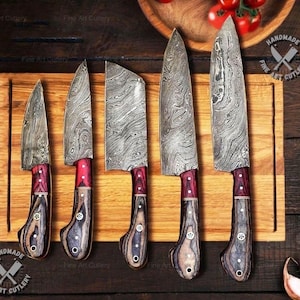 May include: A set of five handmade knives with patterned steel blades and red and wood-toned handles. The knives are displayed on a wooden cutting board. The image includes the text "Fine Art Cutlery" and "HANDMADE FINE ART CUTLERY".