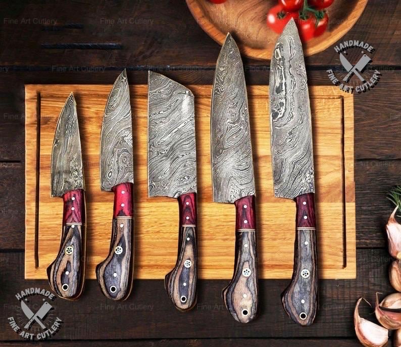 May include: A set of five handmade knives with patterned steel blades and red and wood-toned handles. The knives are displayed on a wooden cutting board. The image includes the text "Fine Art Cutlery" and "HANDMADE FINE ART CUTLERY".