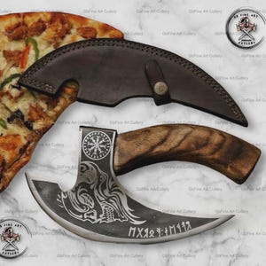 Könnte beinhalten: Ein gebogenes Pizzamesser in Silberfarbe mit Holzgriff und Lederscheide. Die Klinge hat geätzte Designs und Text. Das Messer liegt neben einer Pizzaschnitte. Das Messer ist von Gb Fine Art Cutlery.