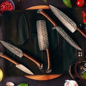 May include: A collection of handcrafted knives with wooden handles and textured blades, displayed on a black leather knife roll. The knives vary in size and shape, with a variety of blade patterns. The scene includes fresh tomatoes, herbs, and spices.