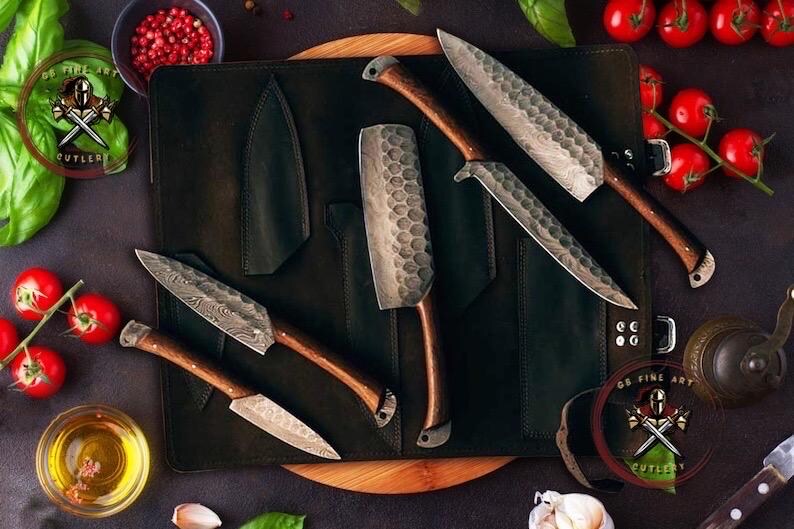 May include: A collection of handcrafted knives with wooden handles and textured blades, displayed on a black leather knife roll. The knives vary in size and shape, with a variety of blade patterns. The scene includes fresh tomatoes, herbs, and spices.