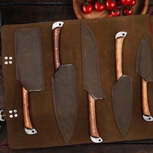 May include: A set of five kitchen knives with wooden handles and leather sheaths, displayed on a brown leather roll. The knives vary in size and shape, with a cleaver and paring knife included. The logo "OF FINE ART CUTLERY" is visible.