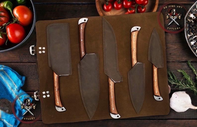 May include: A set of five kitchen knives with wooden handles and leather sheaths, displayed on a brown leather roll. The knives vary in size and shape, with a cleaver and paring knife included. The logo "OF FINE ART CUTLERY" is visible.