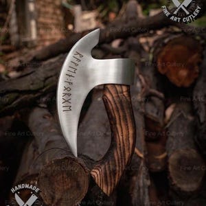 May include: A handmade, curved blade axe with a wooden handle. The silver blade has etched runes. The handle is made of dark, striped wood. The axe is displayed against a backdrop of cut logs. The words "Fine Art Cutlery" and "HANDMADE FINE ART CUTLERY" are visible.