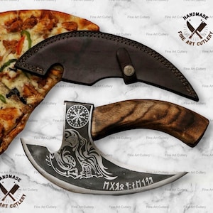May include: A handmade, curved blade knife with a wooden handle and a leather sheath. The blade features etched designs and text. The knife is displayed with a slice of pizza. The text on the knife reads "MXяя : M1198".