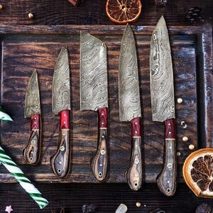 May include: Five Damascus steel knives with wooden handles and red accents are displayed on a wooden tray. A green and white candy cane is in the foreground, with dried orange slices and spices scattered around.