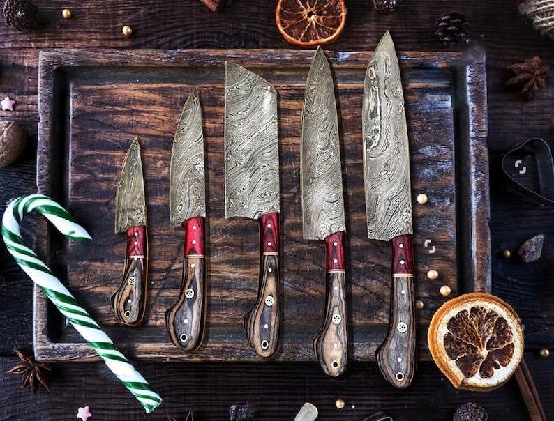 May include: Five Damascus steel knives with wooden handles and red accents are displayed on a wooden tray. A green and white candy cane is in the foreground, with dried orange slices and spices scattered around.