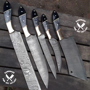 Op de afbeelding: Een set van zes handgemaakte kunstmessen. De messen hebben een uniek patroon. De handvatten zijn zwart en grijs met messing accenten. De set bevat een hakmes en verschillende mesmaten. Het logo "HANDMADE FINE ART CUTLERY" is zichtbaar.