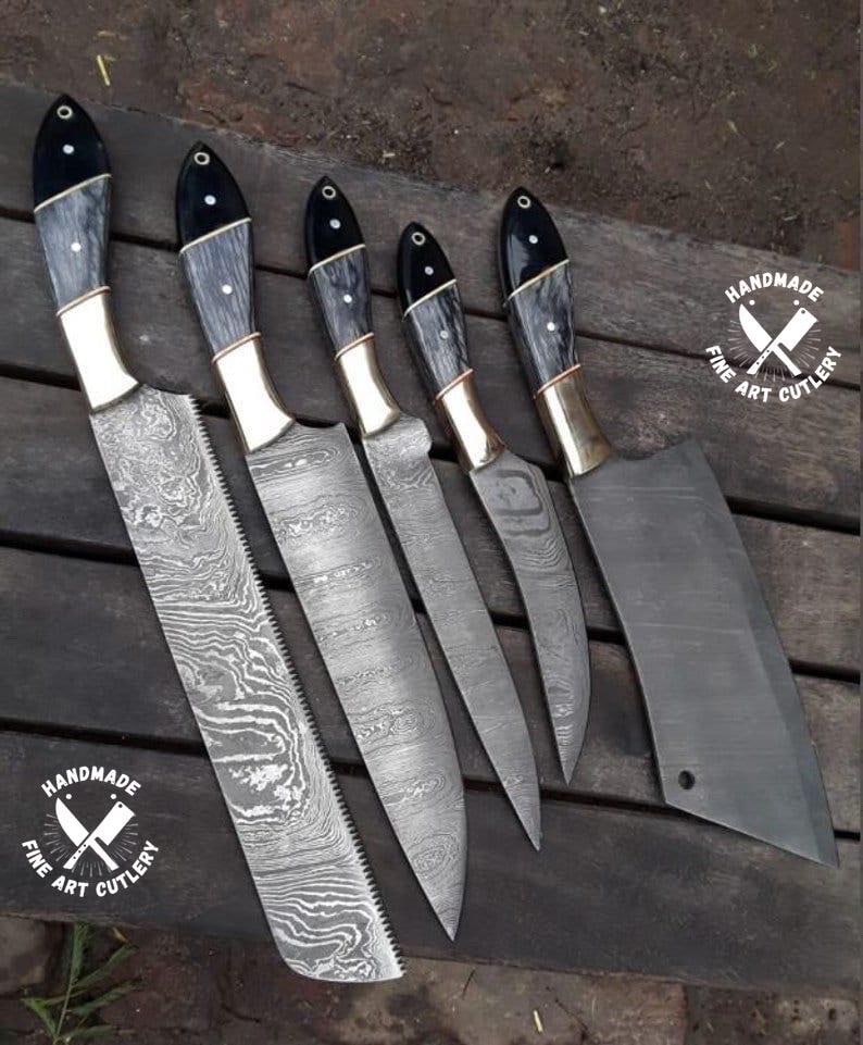 May include: A set of six handmade fine art cutlery knives. The blades have a unique patterned design. The handles are black and gray with brass accents. The set includes a cleaver and various knife sizes. The logo "HANDMADE FINE ART CUTLERY" is visible.