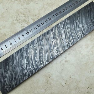 May include: A rectangular steel plate with a distinctive, wavy pattern, alongside a metal ruler. The steel has a dark, marbled appearance with light and dark grey streaks. The ruler shows measurements in centimetres.
