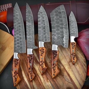 May include: A set of six Damascus steel kitchen knives with wooden handles. The blades have a unique patterned design. The handles are decorated with floral patterns and metal accents. The knives are arranged on a wooden cutting board.