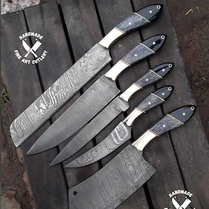 May include: An assortment of handmade fine art cutlery knives with dark wood handles, brass bolsters, and patterned steel blades. The knives are arranged on a dark wooden surface. The image includes a circular logo that says "Handmade Fine Art Cutlery".