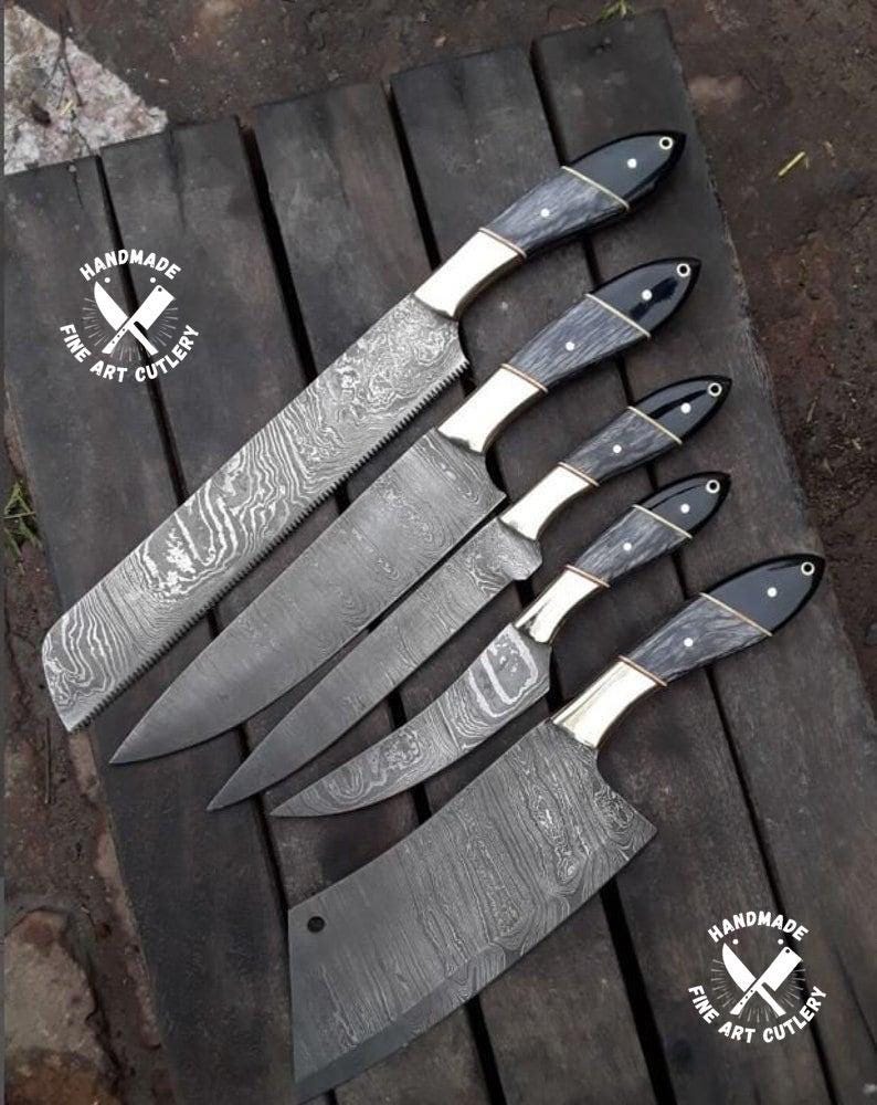 May include: An assortment of handmade fine art cutlery knives with dark wood handles, brass bolsters, and patterned steel blades. The knives are arranged on a dark wooden surface. The image includes a circular logo that says "Handmade Fine Art Cutlery".