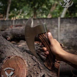May include: A handmade axe with a curved blade and a wooden handle. The blade has etched text. The handle has a dark wood grain pattern. The axe is held over a tree trunk. The image has the text "HANDMADEE FINE ART CUTLERY".