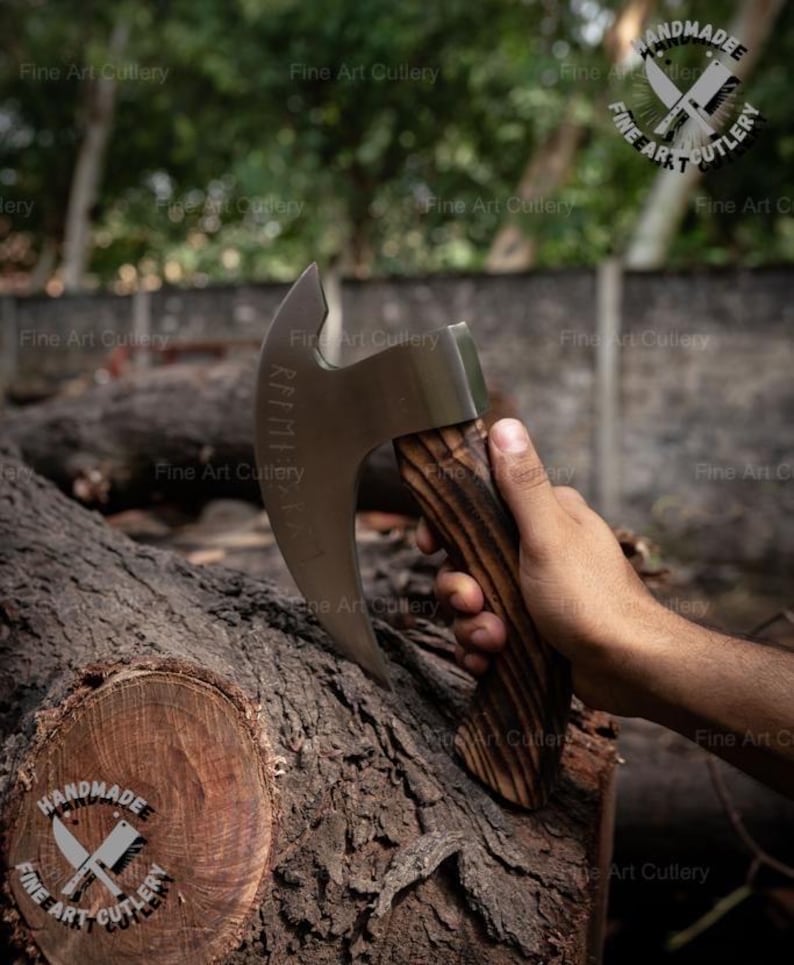May include: A handmade axe with a curved blade and a wooden handle. The blade has etched text. The handle has a dark wood grain pattern. The axe is held over a tree trunk. The image has the text "HANDMADEE FINE ART CUTLERY".