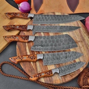 May include: A set of six Damascus steel kitchen knives with wooden handles. The blades have a unique patterned design. The handles are a light brown color with decorative floral accents. The knives are arranged on a wooden cutting board.