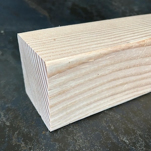 May include: Close-up of a light-colored, rectangular wooden block. The wood grain is visible, running horizontally across the top and vertically on the end. The block is likely for woodworking or construction.