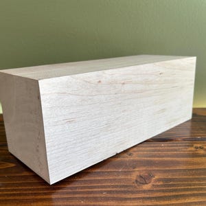 May include: A rectangular block of light-colored, unfinished wood with a visible natural grain pattern. The object has sharp, clean edges and a smooth, matte surface, showcasing the raw texture and pale beige tones of the timber material.