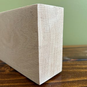 May include: A rectangular block of light-colored, unfinished wood with a visible grain pattern. The piece features a smooth, flat surface on the front and a textured, porous end grain on the side, showcasing the natural light beige tones of the timber.
