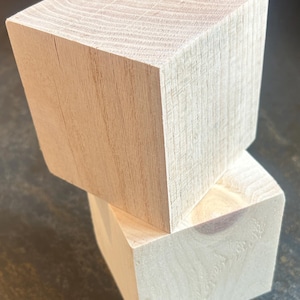 May include: Two unfinished wooden cubes stacked at an angle. The cubes are light-colored with visible wood grain and a natural, unpainted finish. The top cube is slightly angled, revealing the cut edges.