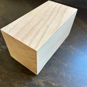 May include: A light-colored, rectangular wooden block with visible grain. The block has a slightly angled side, giving it a trapezoidal shape. The wood appears to be unfinished.