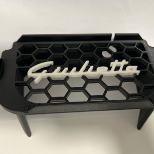 Può includere: Griglia per auto in plastica nera con logo "Giulietta" bianco. La griglia ha un motivo a nido d'ape ed è progettata per essere montata su un'auto.