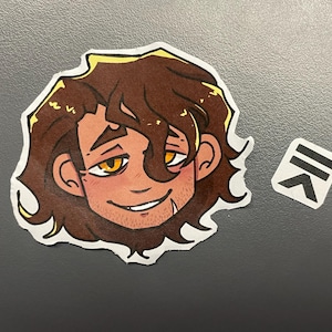 May include: A sticker featuring a cartoon illustration of a smiling person with brown hair and yellow eyes. The sticker has a white border and a small logo sticker is next to it.